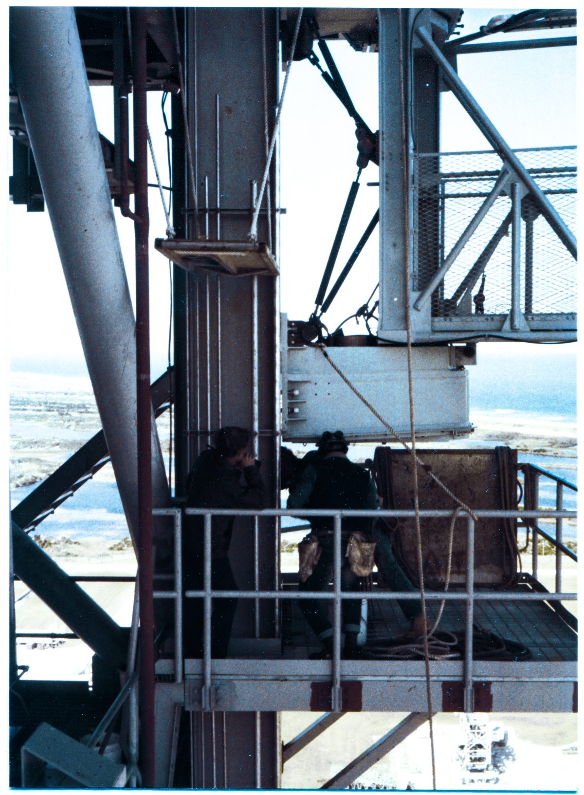 Image 115. Viewed from the GOX Arm Upper Latchback Access Platform, a very judicious choice for exact viewing angle permits us to see the GOX Arm Strongback Columns, and the GOX Arm Lower Hinge Box Connection Plates, from precisely side-on. That the Upper Latchback Access Platform existed where it did on the Fixed Service Structure, high above the Pad Deck at Space Shuttle Launch Complex 39-B, Kennedy Space Center, Florida, was most fortuitous, and our photographer did not squander this opportunity to let us see just how close we are to having the the GOX Arm finish-bolted to the tower. Union Ironworkers working for Ivey Steel Erectors, have at this point gotten the Arm into a position where the holes in the Hinge Box Connection Plates and the holes in the Strongback Columns they attach to are sufficiently lined-up to permit bolts to be inserted through them without damaging the threads, with nuts screwed on to the ends of the bolts, only finger-tight at this time, and not yet fully spun down the bolt threads and properly torqued to complete the attachment process. A knife-edge of daylight is visibly showing between the Strongback and the Hinge Box, and interrupting that knife-edge of daylight, the shanks of the bolts are clearly visible, in-place. The heads of the bolts, on the left side, are obscured from view by a run of conduit, which is attached to the back side of the Strongback Column Flange, but on the other end of the bolts, we can clearly see the nuts in place, as well as the washers which will rest beneath them once they are finish-torqued. Elsewhere on the Upper and Lower Hinge Boxes, out of frame and unseeable in this photograph, work continues with the peculiar combination of overwhelming brute force and a surgeon's delicate finesse which you only get with ironworking, causing the rest of the bolt holes to come into proper alignment, which will then allow for the GOX Arm to at-last get securely and permanently fastened to the FSS, where it can do its job supporting Space Shuttle launches. Photo by James MacLaren.